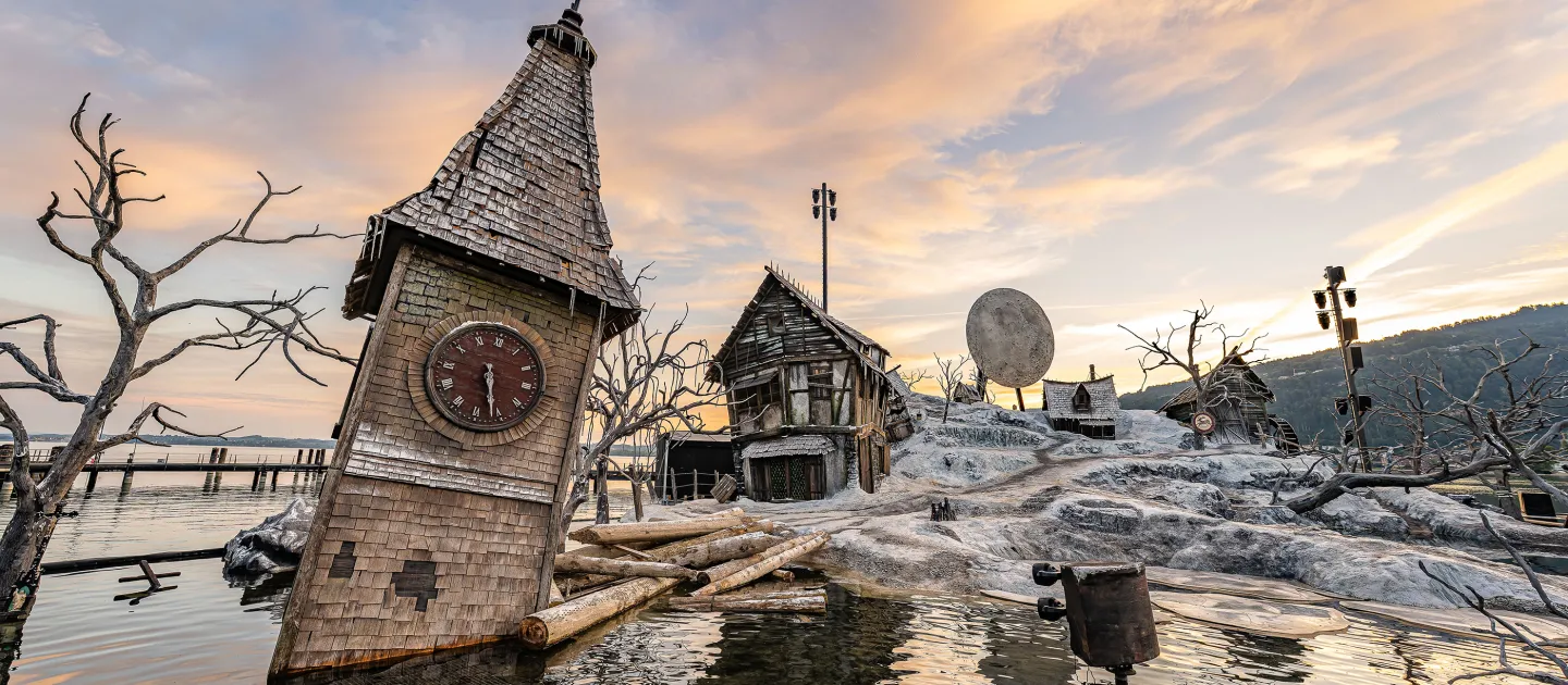 Schief stehender Uhrturm und verfallene Fachwerkhäuser in einer düsteren, wasserüberschwemmten Landschaft mit kahlen Bäumen, Bühnenbild der Oper 'Der Freischütz' bei Sonnenuntergang