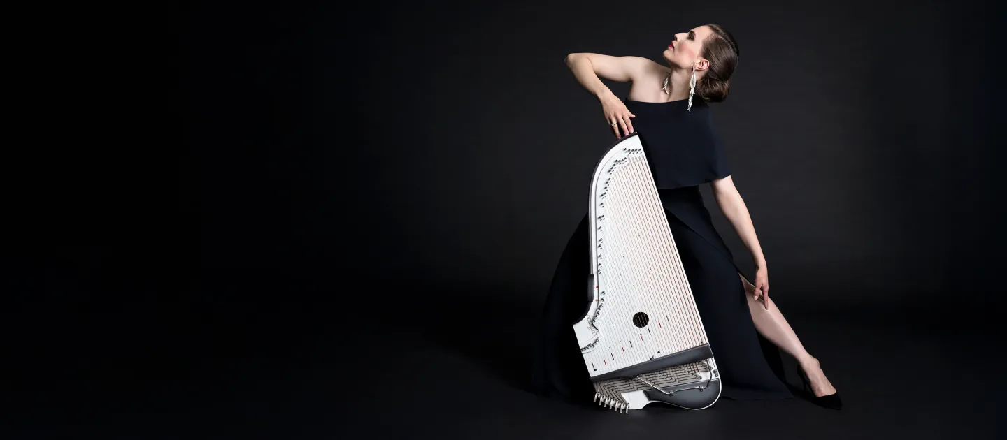Ida Elina in schwarzem Kleid posiert elegant mit einer weißen Kantele vor schwarzem Hintergrund