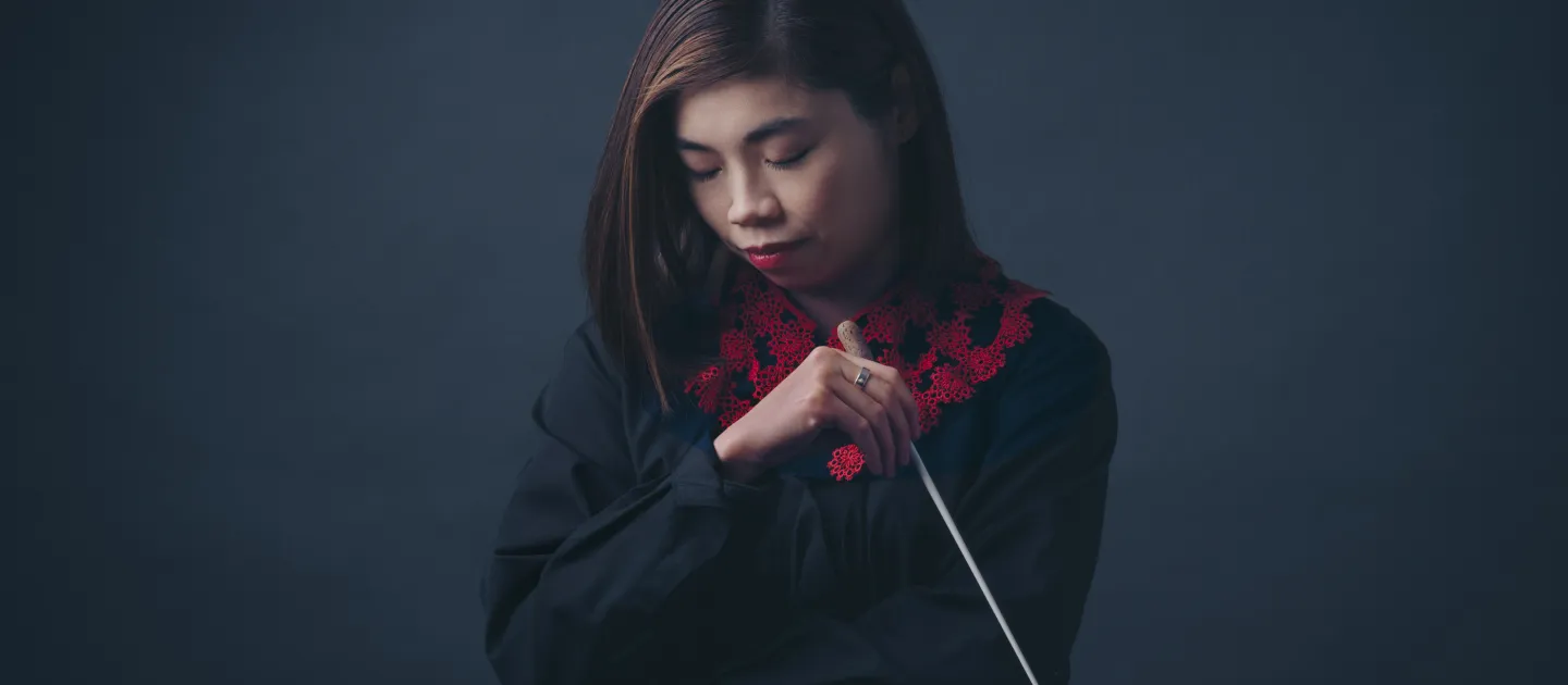 Dirigentin Elim Chan konzentriert mit geschlossenen Augen, hält Taktstock vor dunklem Hintergrund, trägt schwarzes Oberteil mit rotem Muster