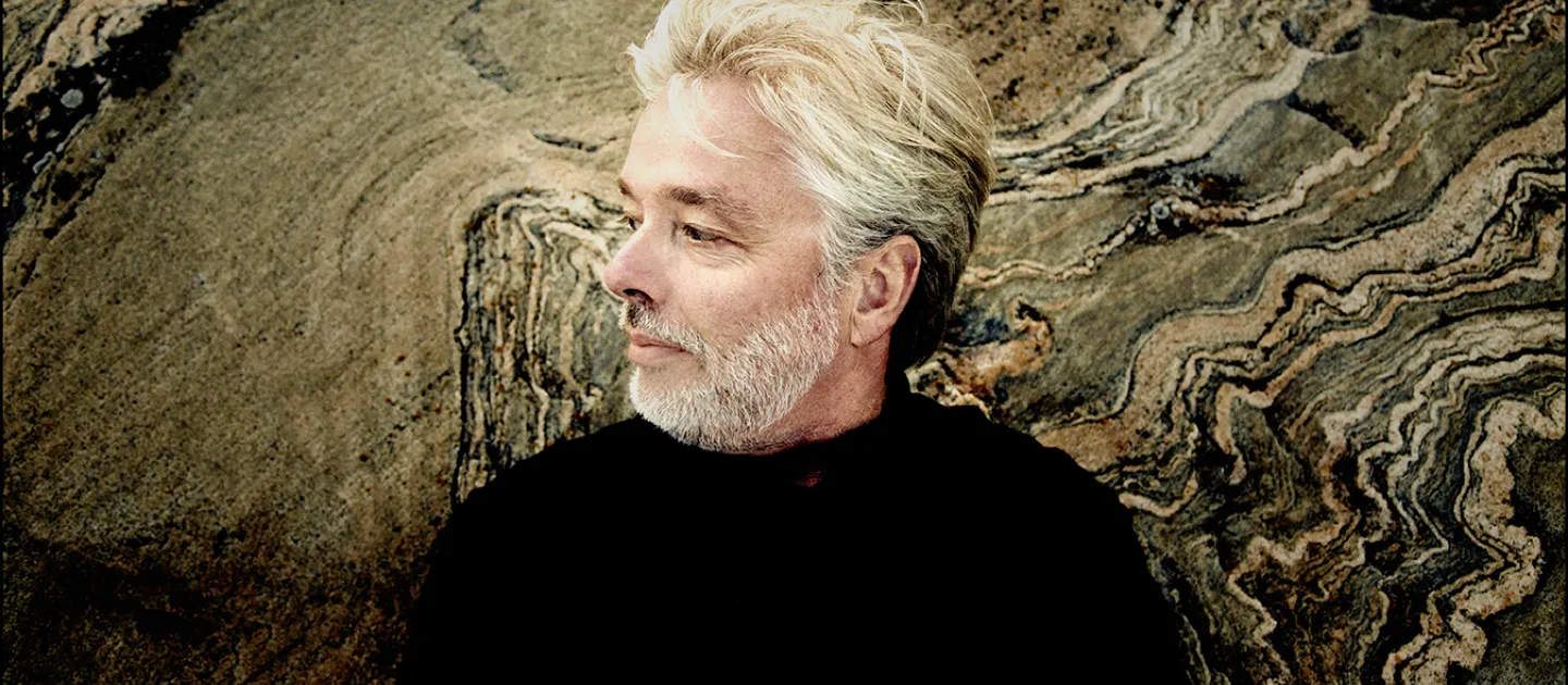 Jukka Pekka Saraste mit grauem Haar und Bart, vor einer strukturierten Felsoberfläche, trägt schwarzen Pullover, Blick nach links