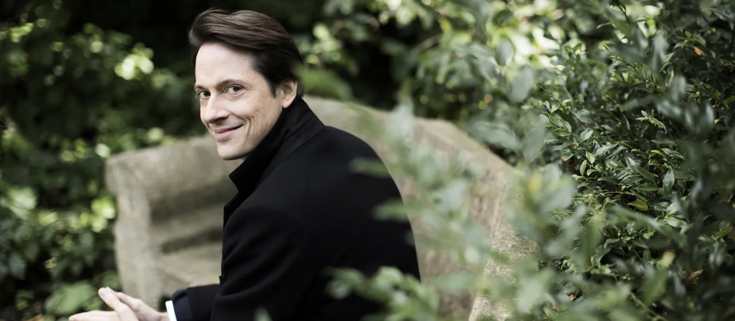 Dirigent Leo McFall sitzt auf einer Steinbank im Grünen, trägt einen schwarzen Mantel und lächelt leicht zur Seite