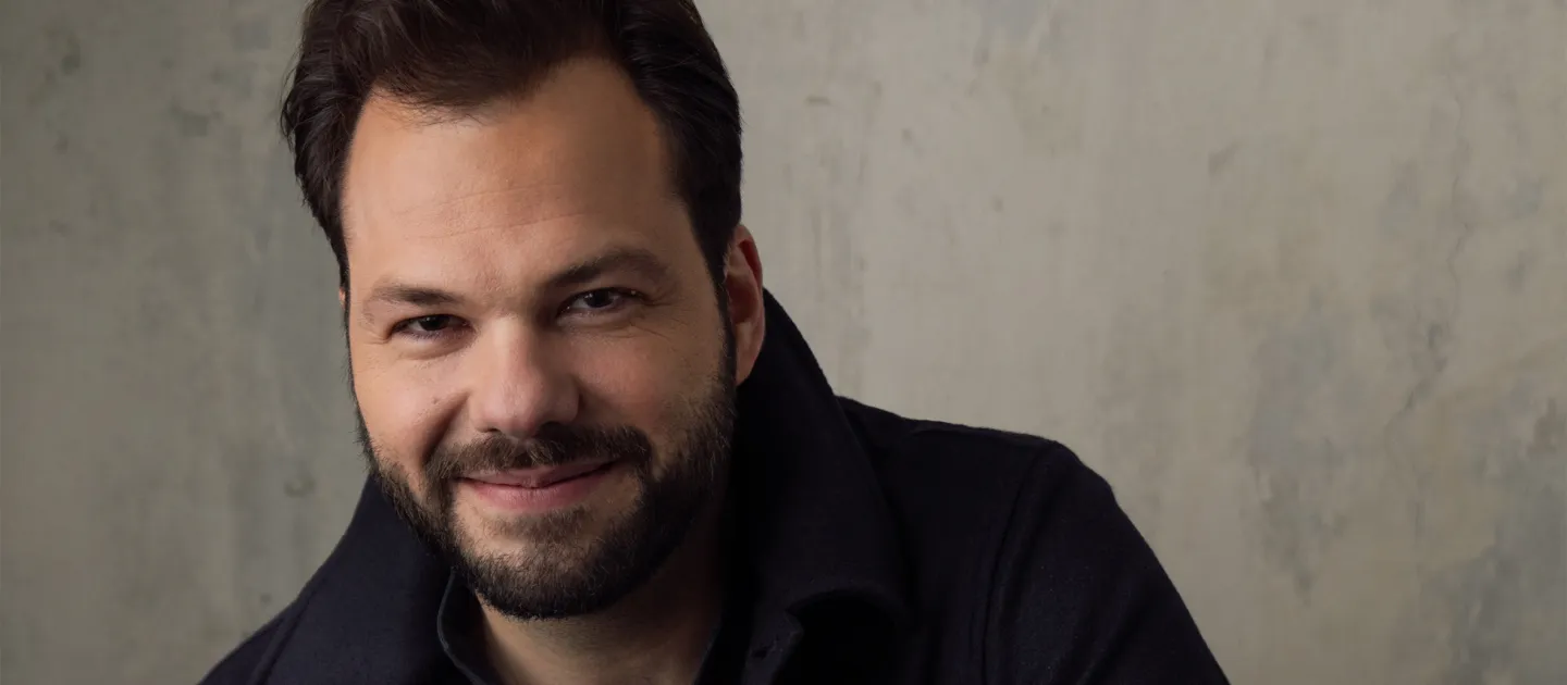 Porträt von Dirigent Petr Popelka mit kurzem dunklem Haar und Bart, lächelnd, vor hellem Hintergrund