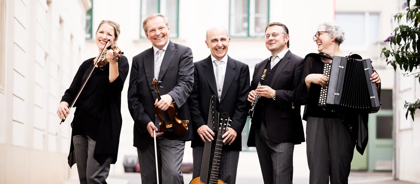 Fünf Musiker in schwarzer Kleidung stehen lachend mit ihren Instrumenten auf einer Straße, darunter Geige, Gitarre, Klarinette und Akkordeon.
