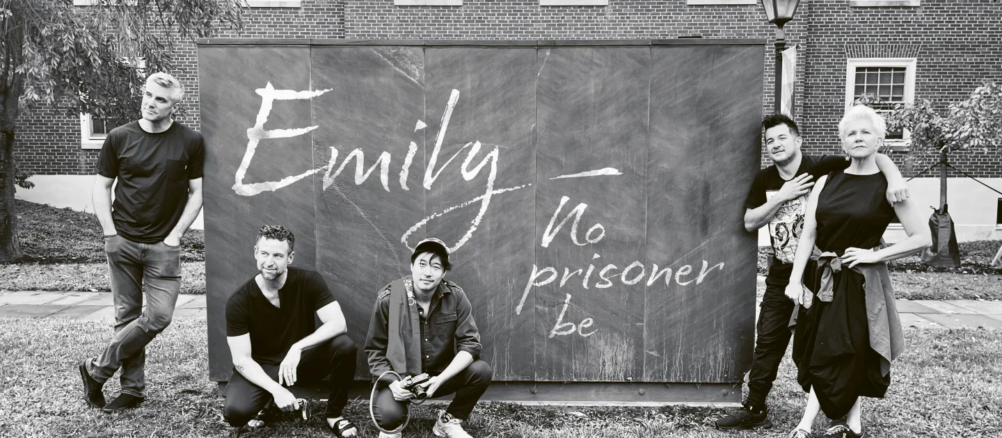 Fünf Personen vor einer Tafel mit der Aufschrift 'Emily - no prisoner be', Kevin Puts, das musikalische Trio time for Three und Joyce DiDonato, entspannt im Freien vor einem Backsteingebäude