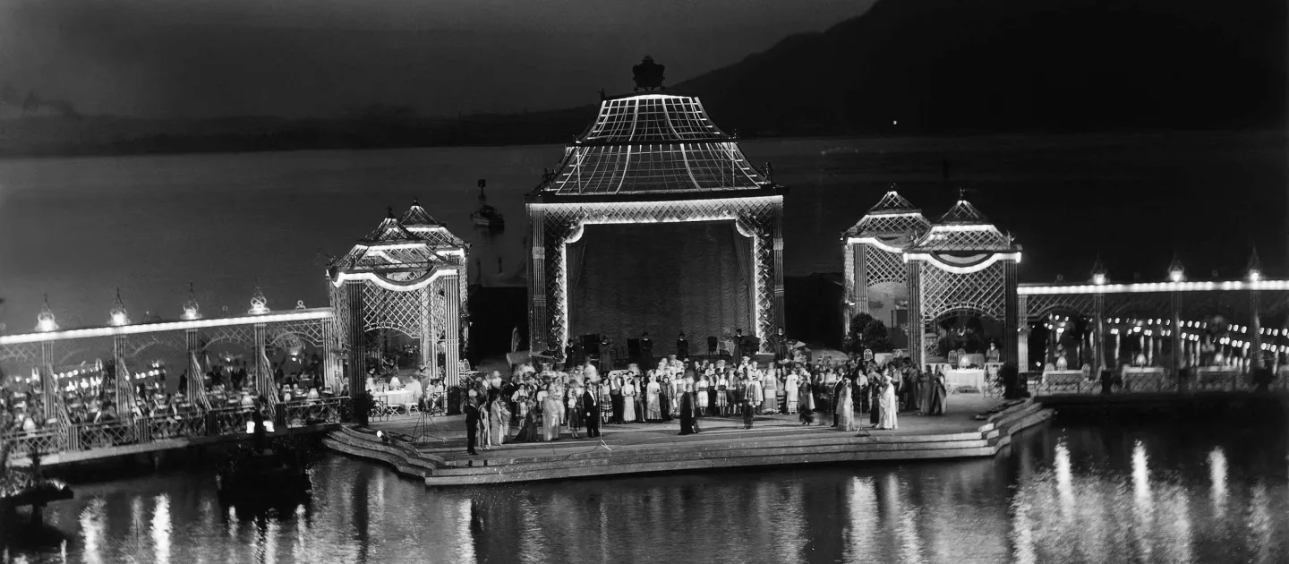 Szene aus Fledermaus, 1954 auf der Seebühne, viele Menschen, im Mittelpunkt befindet sich ein großer Pavillon, links und rechts stehen je zwei weitere, kleinere Pavillons, von denen je ein Steg abgeht