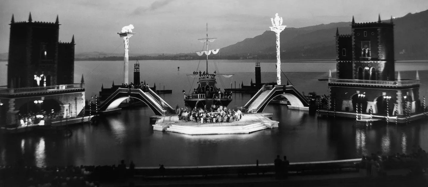 Szene aus "Eine Nacht in Venedig", 1955 auf der Seebühne, im Mittelpunkt befindet sich eine Insel mit einer Menschengruppe, dahinter liegt ein großes Schiff, links und rechts der Insel führt jeweils eine Brücke zu zwei weiteren Inseln, auf denen je ein zweistöckiges Gebäude steht