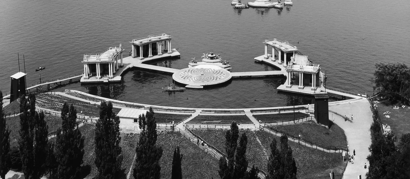 Szene aus "Der Bettelstudent", 1956 auf der Seebühne, im Mittelpunkt befindet sich eine runde Insel mit einem Boden in überwiegend schwarz-weißem Schachbrettmuster, am hinteren Bereich schließt sich ein schmaler Anbau mit vier Skulpturen und einem zentralen Brunnen an, links und rechts führt jeweils ein Steg zu zwei weiteren Inseln, auf denen je zwei tempelartige Gebäude mit Säulen stehen
