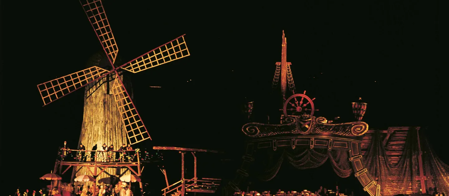 Szene aus "Zar und Zimmermann", 1957 auf der Seebühne, links befindet sich eine Plattform mit einer Windmühle aus Holz und einer Menschengruppe, an deren rechten Seite schließt über eine Brücke ein zweistöckiges Schiff an, im Untergeschoss des Schiffs befindet sich eine große Menschengruppe
