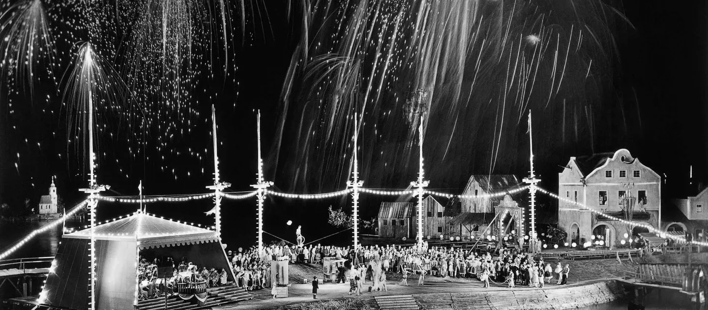 Szene aus "Die verkaufte Braut", 1958 auf der Seebühne, links befindet sich ein Festzelt mit einer Menschengruppe, rechts daneben liegt ein Festplatz, gefüllt mit Menschen, im rechten Hintergrund ist eine Häuserreihung zu sehen 