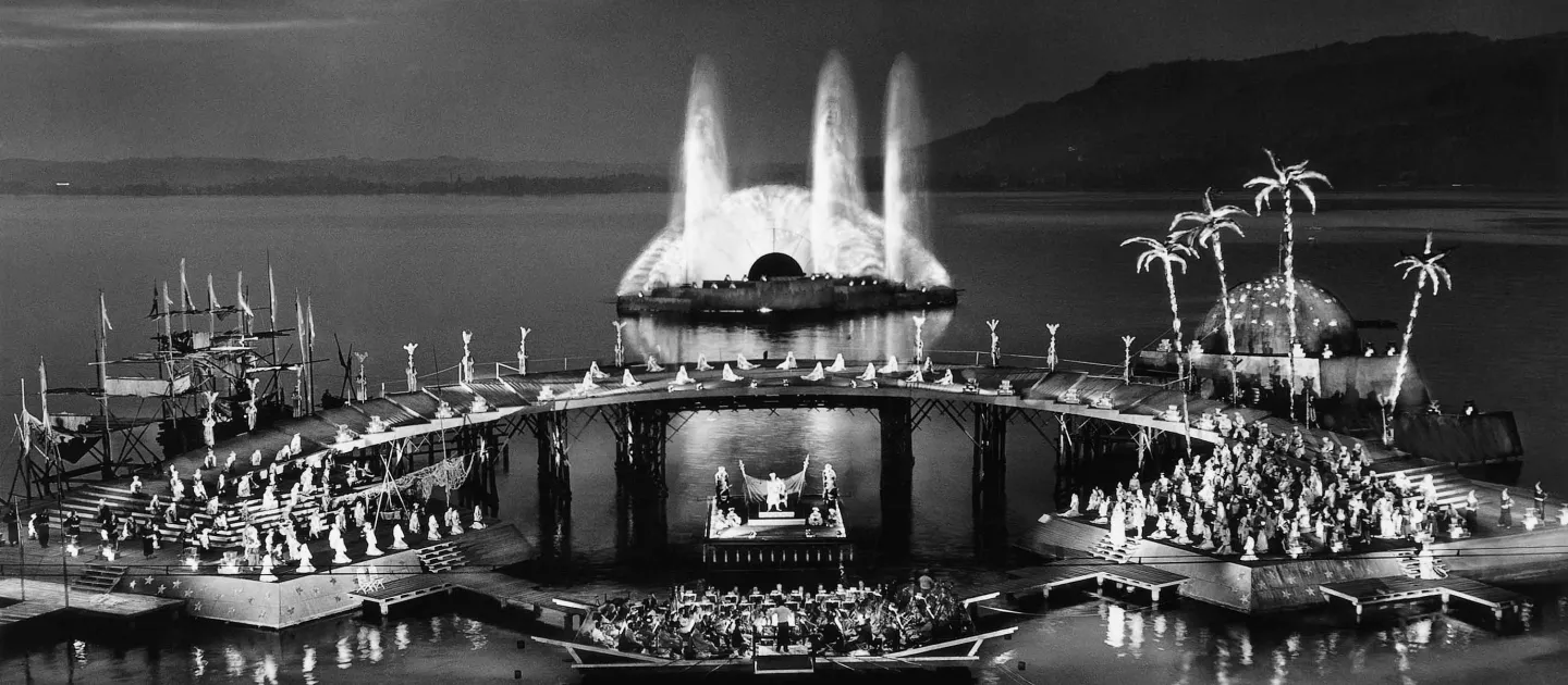 Szene aus "Tausendundeine Nacht", 1959 auf der Seebühne, im Vordergrund befindet sich mittig eine kleine Plattform mit einer kleinen Gruppe von Menschen, links und rechts daneben liegen je eine größere Plattform mit einer großen Menschenmenge, die durch einen höher gelegenen Steg miteinander verbunden sind, hinter dem Steg sind auf der linken Seite Schiffsmasten zu sehen, auf der rechten Seite ein Gebäude und Palmen, im Hintergrund ist eine weitere Plattform mit großem Wasserspiel zu sehen