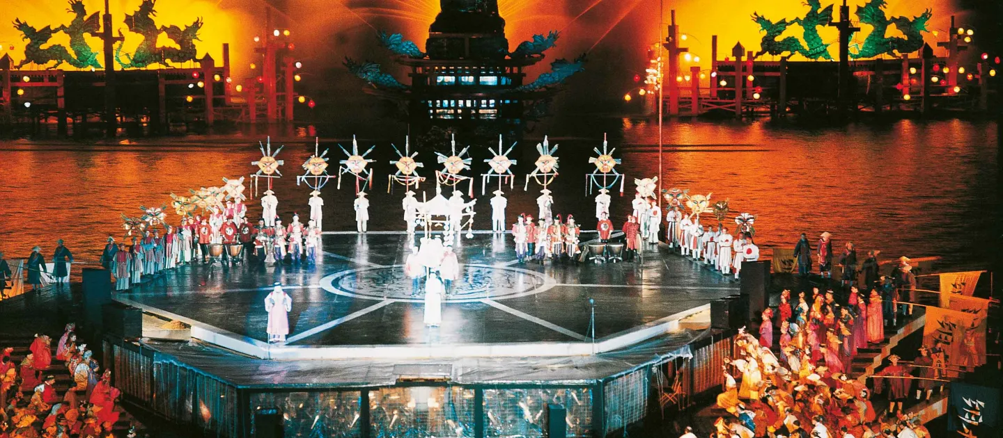 Szene aus "Das Land des Lächelns", 1964 auf der Seebühne, mittig befindet sich eine erhöhte Plattform mit einer Menschengruppe in traditionell chinesischer Kleidung, die hintere Reihe hält Bühnenbildelemente in die Höhe, links und rechts ist je eine Plattform nach unten versetzt mit einer großen Menge an Darstellenden in traditionell chinesischer Kleidung, im Hintergrund sind drei chinesische Skulpturen durch Wasserspiele sowie rotes Licht in Szene gesetzt 