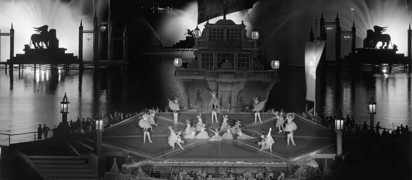 Szene aus "Eine Nacht in Venedig", 1965 auf der Seebühne, mittig befindet sich eine Plattform in Form eines Hexagon mit verschiedenen Balletttänzer:innen, an der Rückseite ist ein mehrstöckiges historisch-aussehendes Schiff angeschlossen, im Hintergrund ist links und rechts eine weitere Plattform mit je einer Silhouette eines geflügelten Löwen (Markuslöwe) und mittig eine Plattform mit einem geflügelten Pferd (Pegasus) zu sehen