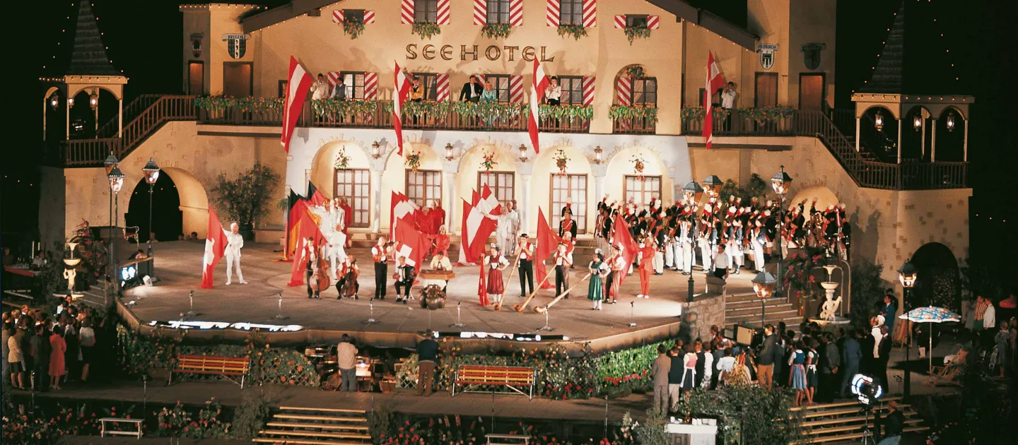 Szene aus "Hochzeit am Bodensee", 1969 auf der Seebühne, eine eckig gestaltete Plattform, auf der ein zweistöckiges Gebäude mit der Aufschrift "Seehotel" steht, zahlreiche Darstellende, teils in Uniform, befinden sich auf der Bühne, viele österreichische Nationalflaggen schmücken das Bühnenbild 