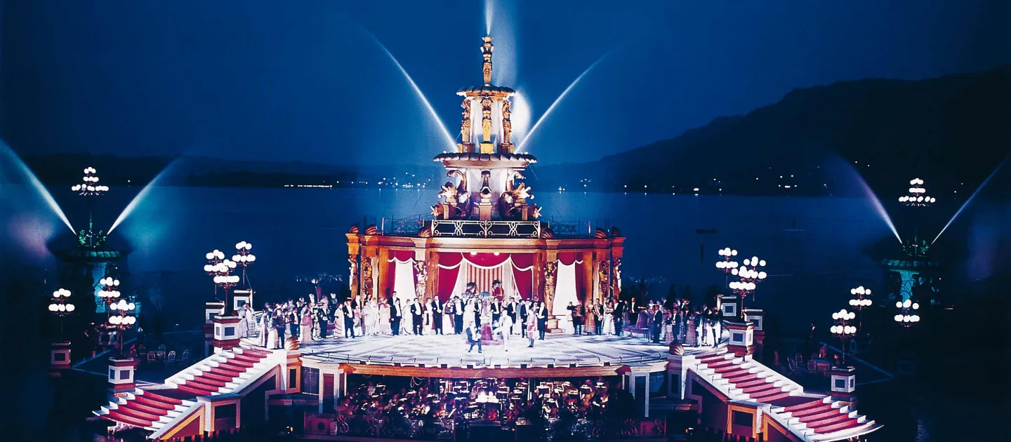 Szene aus "Die Fledermaus", 1970 auf der Seebühne, eine eckig gestaltete Plattform mit einem Gebäude, das über mehrere Höhenniveaus verfügt, die sich nach oben hin verjüngen, von der Plattform führen links und rechts je eine Treppe hinab ins Wasser, das Bühnenbild ist in Rot und Weiß gehalten, das Gebäude ist mit ornamentalen/figuralen Verzierungen und Vorhängen geschmückt