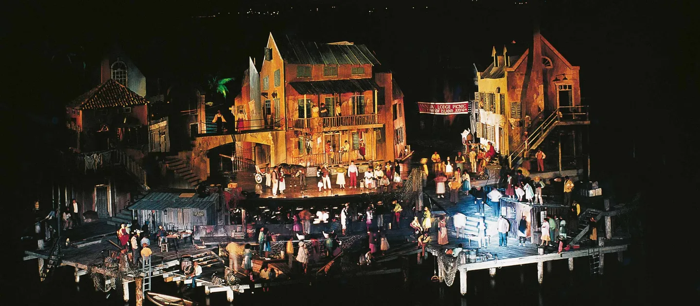 Szene aus "Porgy and Bess", 1971 auf der Seebühne, eine Plattform wird seitlich von zwei kleineren, tiefer liegenden Plattformen flankiert, die über Treppen zugänglich sind, im Zentrum steht ein dreistöckiges Haus, das in rötlichem Licht angestrahlt wird; rechts und links davon befindet sich jeweils ein weiteres Haus, zahlreiche Menschen sind auf der Bühne verteilt