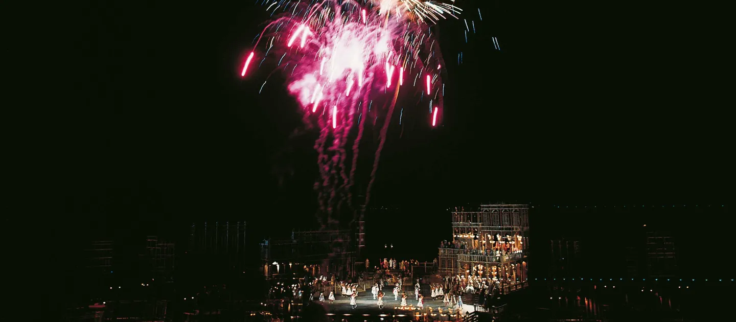 Szene aus "Eine Nacht in Venedig", 1975 auf der Seebühne, dreistöckiges Gebäude, viele Darstellende in Kostümen, am Himmel ist ein buntes Feuerwerk über dem Bühnenbild