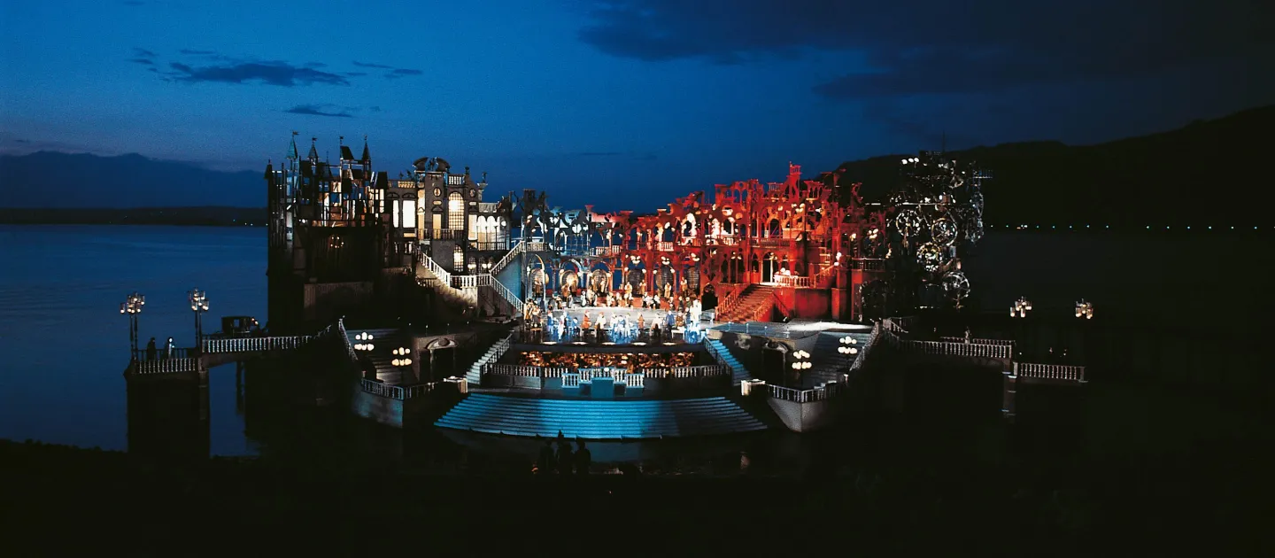 Szene aus "Hoffmanns Erzählungen", 1976 auf der Seebühne, eine Plattform mit unterschiedlichen Höhenniveaus, verbunden durch Treppen, auf der obersten Ebene steht eine bühnenbreite Häuserreihe mit Säulenverzierungen, viele kostümierte Darstellende