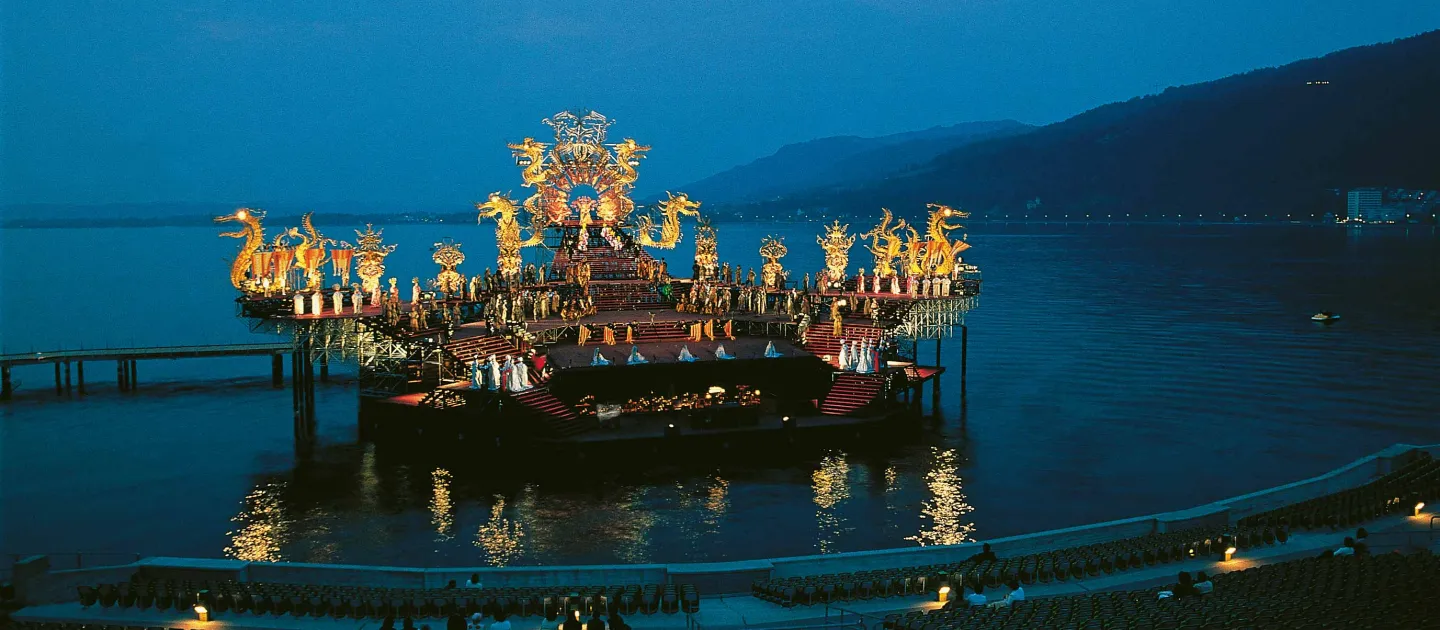 Szene aus "Turandot", 1979 auf der Seebühne, tribünenartiges Bühnenbild mit kostümierten Darstellenden in verschiedenen Ebenen, am Bühnenrand verlaufen goldfarbene Verzierungen mit Drachenfiguren