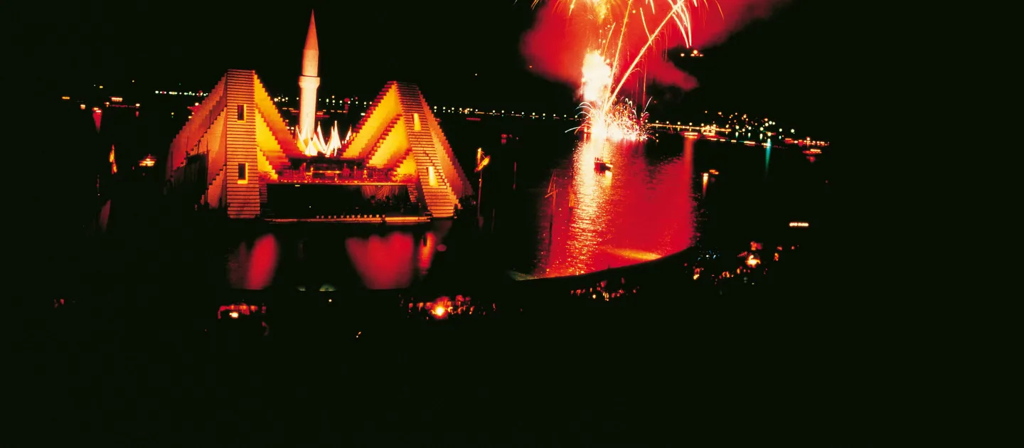 Szene aus "Die Entführung aus dem Serail", 1980 auf der Seebühne, rot beleuchtetes Bühnenbild mit emporragendem Turm, rechts davon ein Feuerwerk über dem Wasser