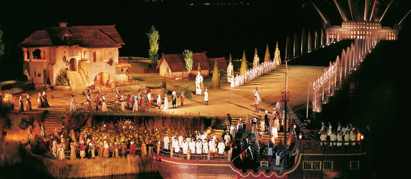 Szene aus "Der Zigeunerbaron", 1982 auf der Seebühne, auf der linken Seite befindet sich ein großes Haus, viele Darstellende in historischen Kostümen, ein Schiff im Wasser im Vordergrund, nahe an der Bühne, im Hintergrund beleuchtete Fontänen, die zu einer beleuchteten Gebäudesilhouette führen
