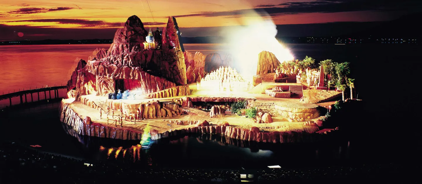 Szene aus "Die Zauberflöte", 1985 auf der Seebühne, Felsenlandschaft mit einzelnen Bäumen, ein größerer Berg befindet sich auf der linken Seite des Bühnenbilds, die Darstellenden sind mittig aufgereiht und hell angeleuchtet 
