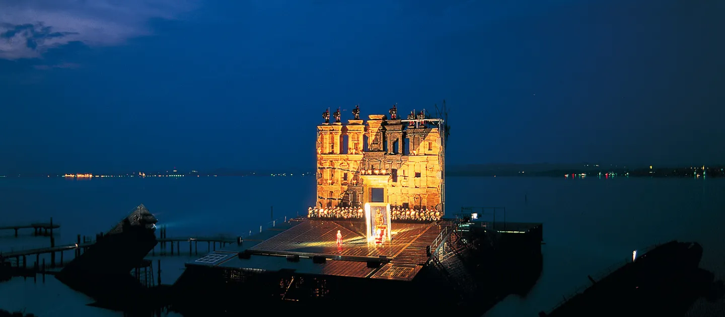 Szene aus "Nabucco", 1994 auf der Seebühne, große Gebäudefassade im Hintergrund, viele kostümierte Darstellende in einer reihe davor, auf der freien Fläche im Vordergrund befindet sich ein großes Reliefbild mit Abbild eines Menschen 