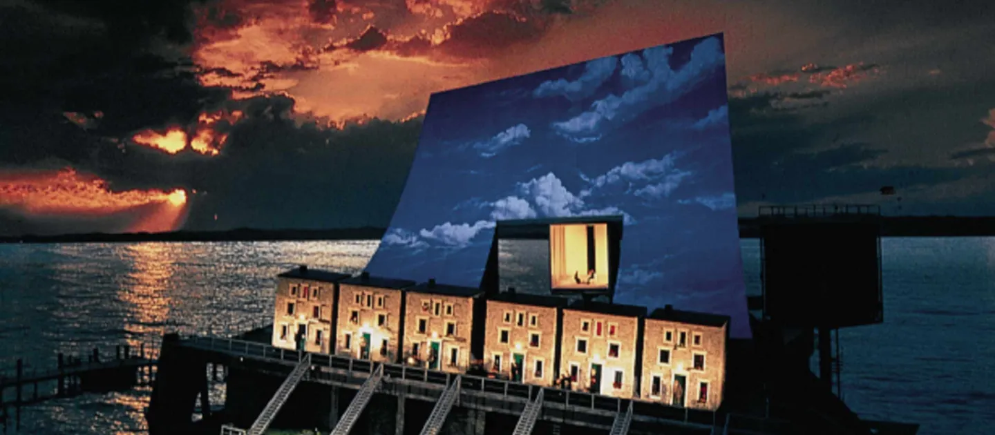 Szene aus "Fidelio", 1995 auf der Seebühne, sechs Häuserfassaden mit Treppen zu einer tieferliegenden Ebene, im Hintergrund großes Wolkenbild mit beleuchteter viereckiger Öffnung