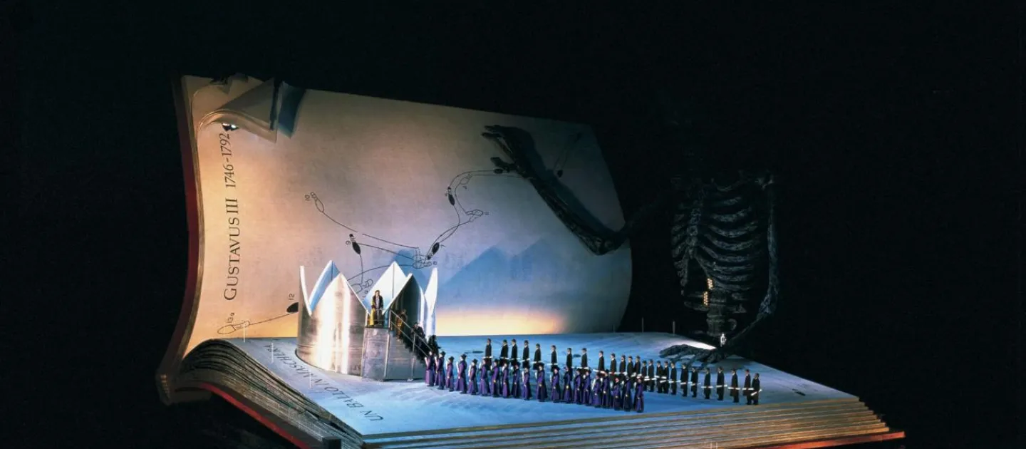 Szene aus "Ein Maskenball", 1999 auf der Seebühne, riesiges aufgeschlagenes Buch als Bühne mit einer großen silberfarbenen Krone links von der Bühne und einer großen Skelettfigur im Hintergrund, zwei Reihen kostümierter Darstellenden in Richtung der Krone 