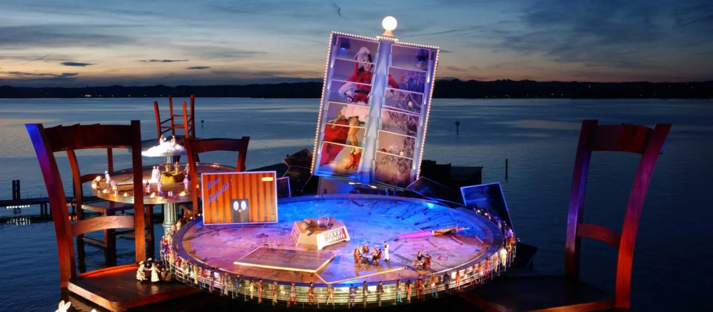 Szene aus "La Bohème", 2001 auf der Seebühne, großer Tisch als Bühnenfläche mit zwei überdimensionierten Stühlen im Wasser, im Hintergrund ein Kartenhalter mit Postkarten, die gemeinsam ein Gesamtbild ergeben und ein weiterer Tisch mit Stühlen, einige kostümierte Darstellende