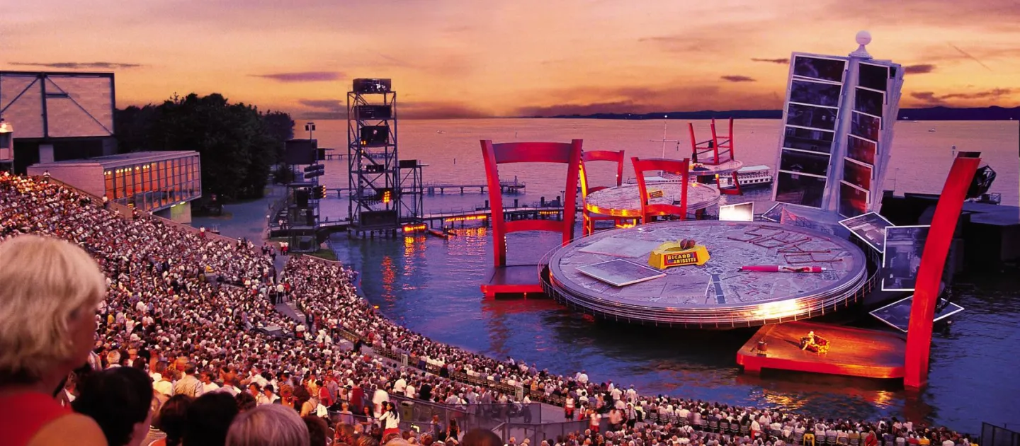 Szene aus "La Bohème", 2002 auf der Seebühne, großer Tisch als Bühnenfläche mit zwei überdimensionierten Stühlen im Wasser, im Hintergrund ein Kartenhalter mit Postkarten, die gemeinsam ein Gesamtbild ergeben und ein weiterer Tisch mit Stühlen
