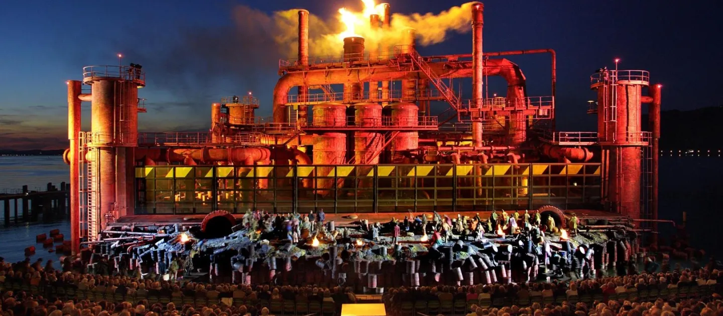 Szene aus "Der Troubadour", 2005 auf der Seebühne, Industrieanlage mit rauchenden Schornsteinen, einige kostümierte Darstellende im Vordergrund