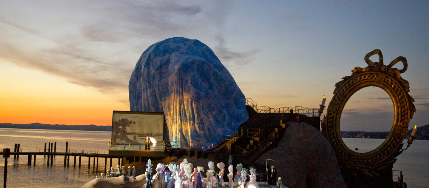 Szene aus "André Chénier", 2011 auf der Seebühne, große Hände umklammern die Seiten der Bühnenfläche, ein goldener Spiegelrahmen im Hintergrund und Figuren in historischen Kostümen auf der Bühnenfläche