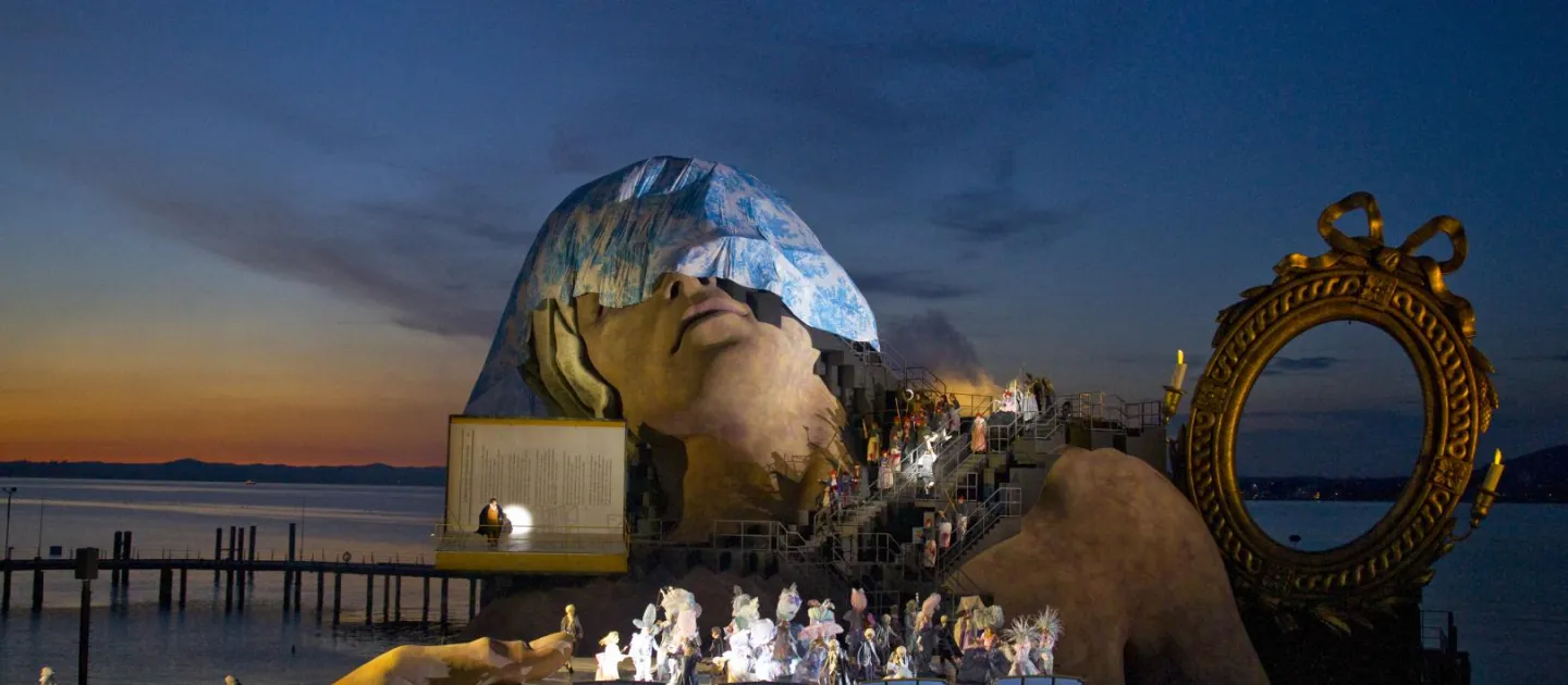 Szene aus "André Chénier", 2012 auf der Seebühne, große Hände umklammern die Seiten der Bühnenfläche, ein goldener Spiegelrahmen und einer riesiger mit einem Tuch bedeckter Kopf im Hintergrund und Figuren in historischen Kostümen auf der Bühnenfläche