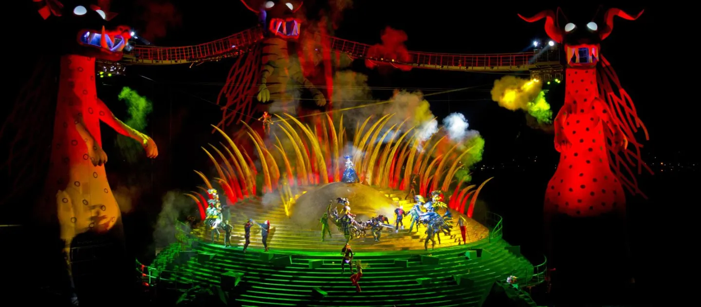 Szene aus "Die Zauberflöte", 2013 auf der Seebühne, im Hintergrund große rot beleuchtete Figuren mit Hörnern und durch Brücken verbunden, mittig eine grün-gelb beleuchtete runde Bühnenplattform, Rauch und mehrere kostümierte Darstellende im Vordergrund