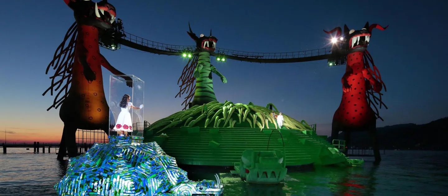 Szene aus "Die Zauberflöte", 2014 auf der Seebühne, drei große Drachenfiguren, durch Brücken verbunden, zwei Inseln im Wasser, im Vordergrund eine Person in einem transparenten Kasten