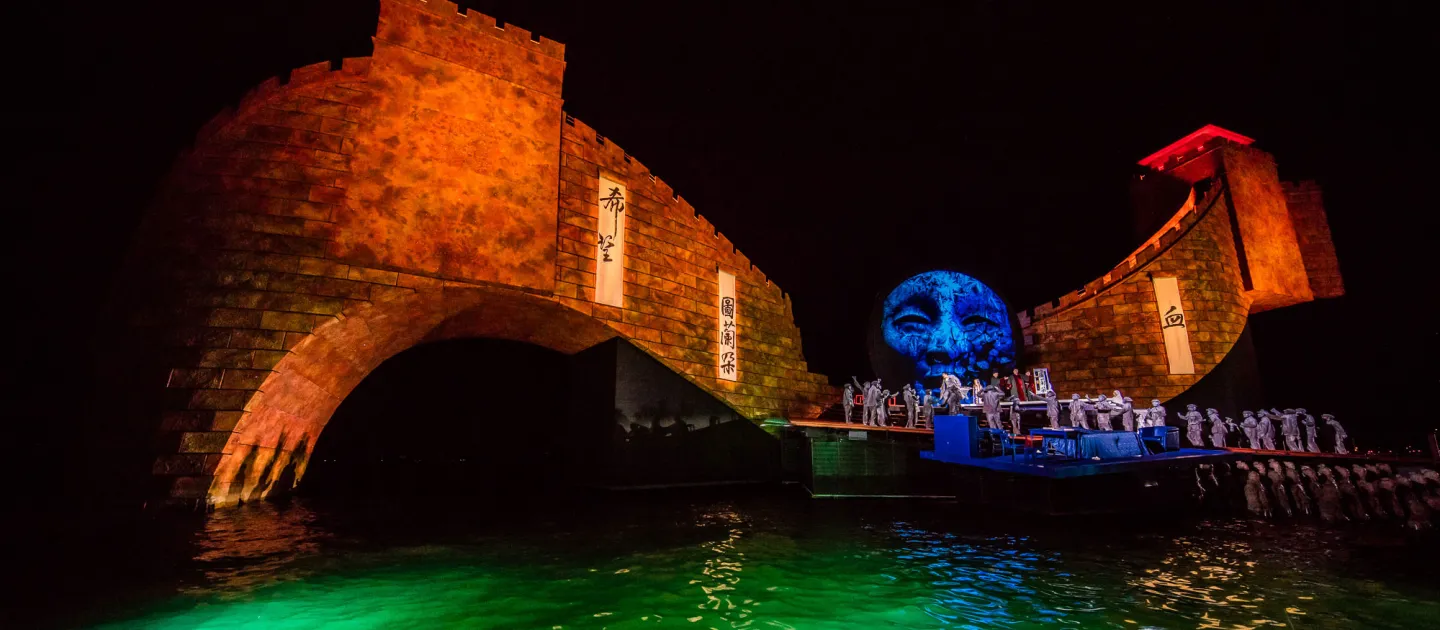 Szene aus "Turandot", 2015 auf der Seebühne, gebogener Steinbogen und großes blaues Gesicht im Hintergrund, im Vordergrund mehrere kostümierte Darstellende