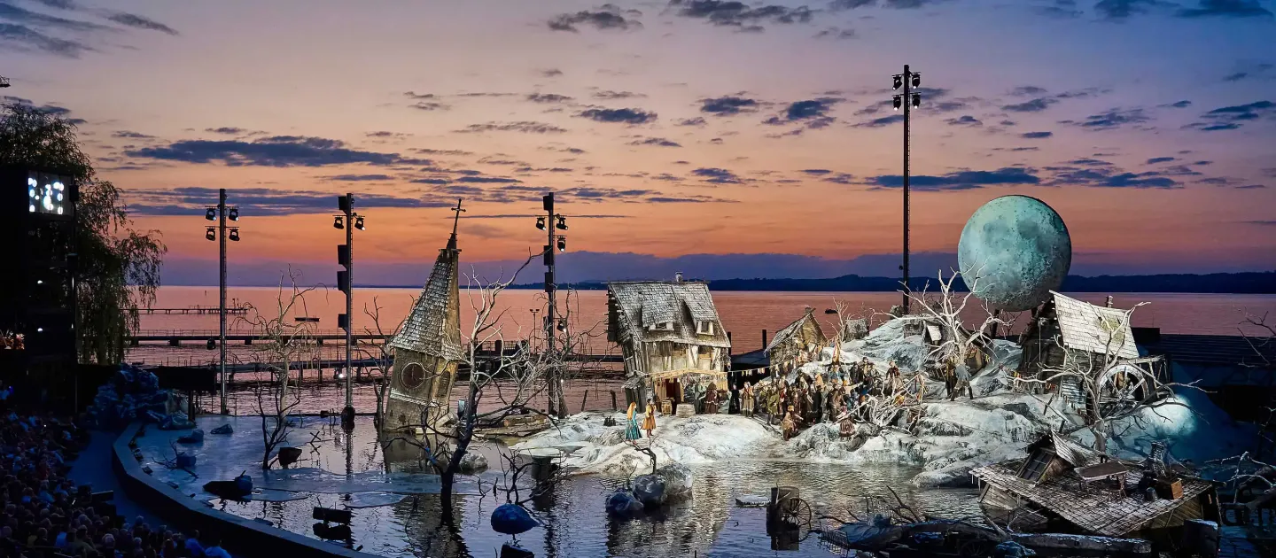 Kulisse "Der Freischütz" - Schneelandschaft überflutet, windschiefe Häuser, ein Kirchturm ragt aus dem Wasser, dramatischer Sonnenuntergang im Hintergrund in orange bis blau