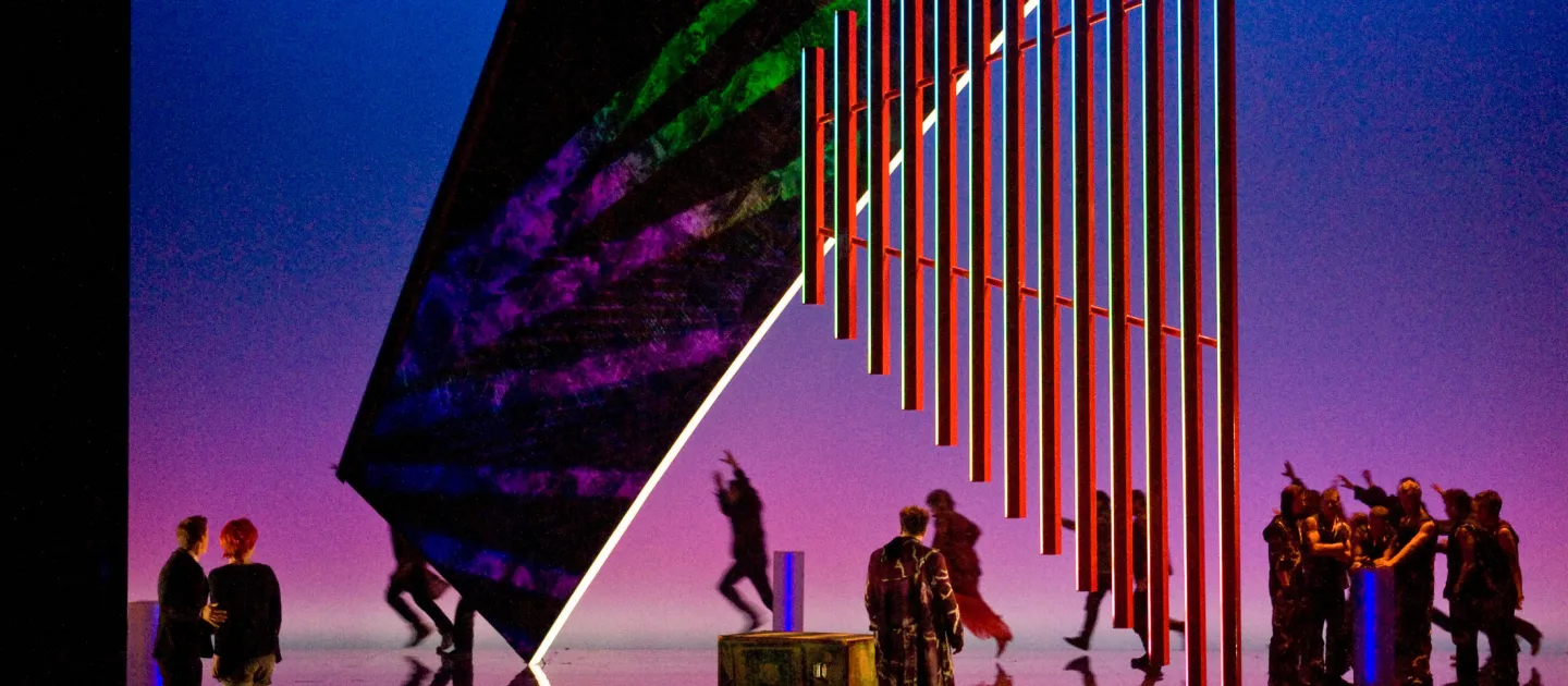 Szene aus "Achterbahn", 2011 im Festspielhaus, mehrere Darstellende tanzen, eine große Struktur hängt von der Decke, verspiegelter Boden, blau-pinker Hintergrund 