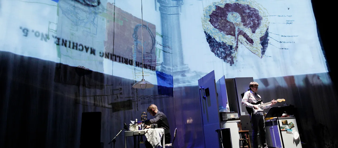 Szene aus "Home Work", 2011 im Festspielhaus, zwei Personen, eine am Tisch, die andere spielt Gitarre, Projektionen auf Leinwänden im Hintergrund, seitliche Aufnahme