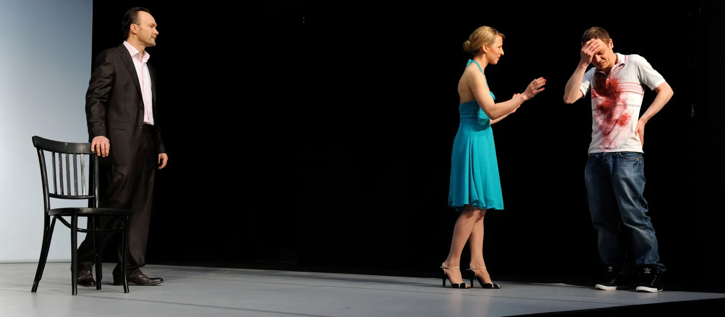 Szene aus "Waisen", 2011 im Theater Kosmos, drei kostümierte Darstellende, links ein Mann in schwarzem Anzug und weißem Hemd neben schwarzem Stuhl, rechts eine Frau in türkisem Kleid gestikuliert zu einem Mann mit kunstblutbeflecktem weißem T-Shirt