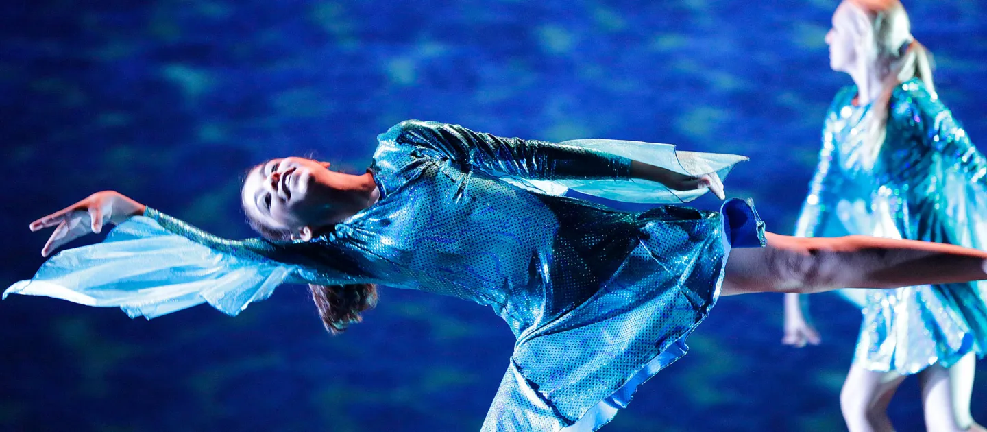 Szene aus "Panta rhei – alles fließt!", 2012 im Festspielhaus, im Vordergrund steht eine blau kostümierte Darstellerin auf einem Bein, das andere zur Seite gestreckt, der Oberkörper nach vorner gelehnt, rechter Arm ausgestreckt, im Hintergrund eine weitere Person im blauen Kostüm