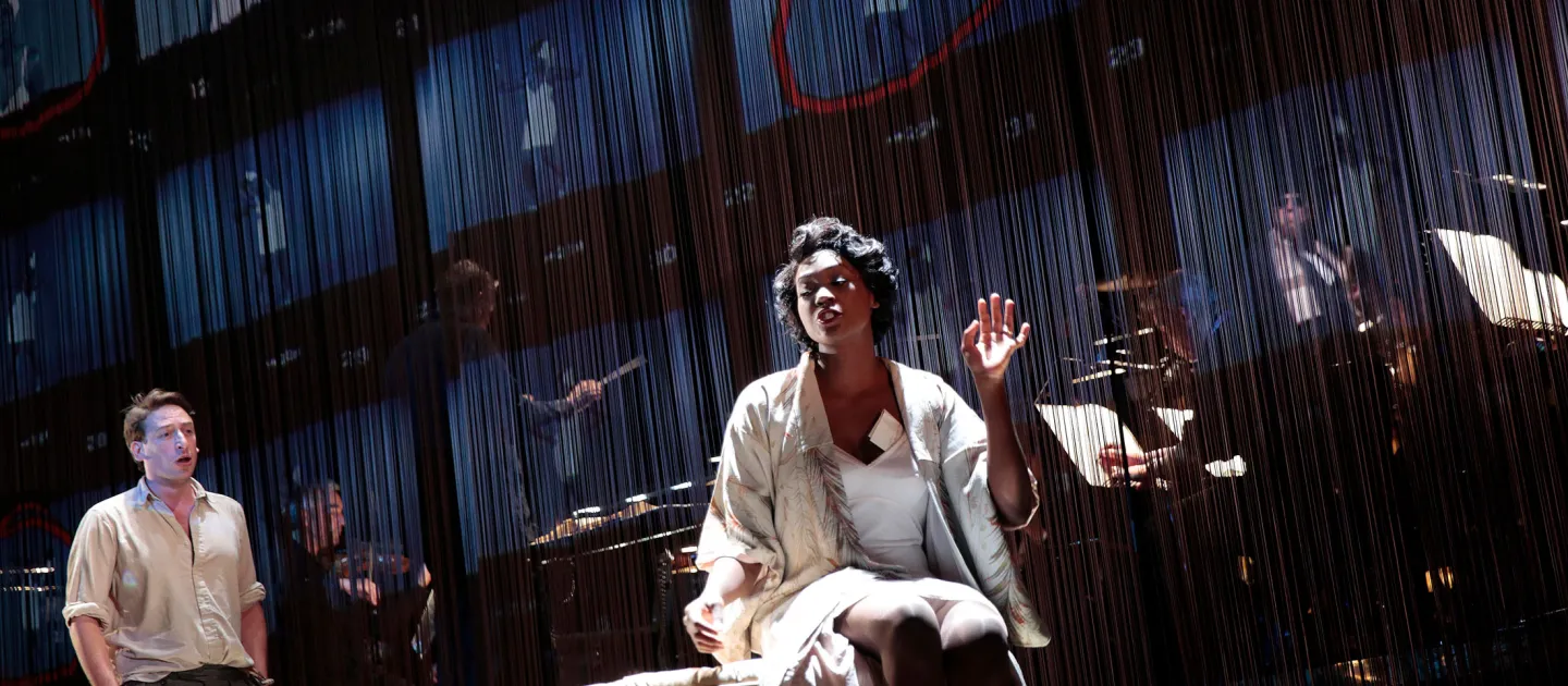 Szene aus "American Lulu", 2013 im Festspielhaus, zwei Darstellende, im Vordergrund eine Darstellerin sitzt und hebt eine Hand, links ein weiterer Darsteller, im Hintergrund von der Decke hängende Fäden und Musiker:innen