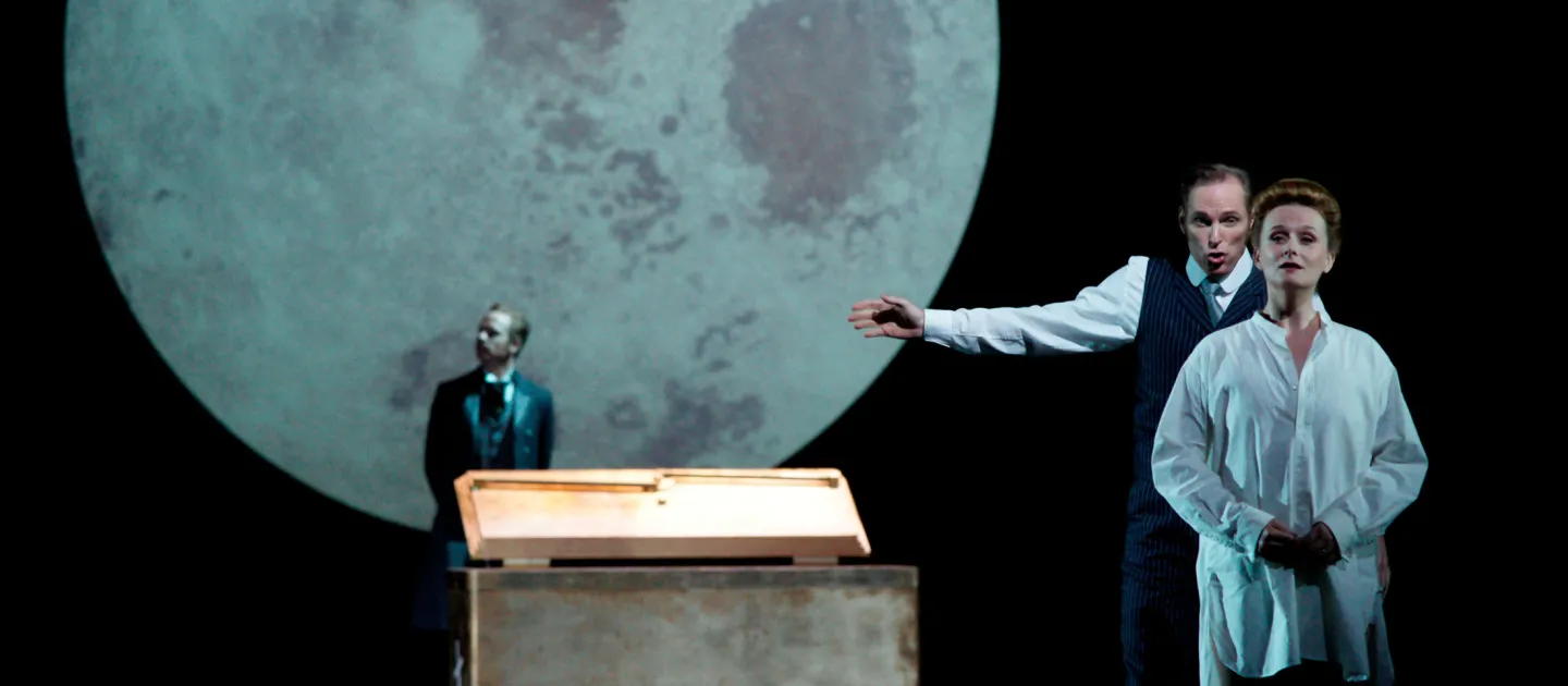 Szene aus "Der Kaufmann von Venedig", 2013 im Festspielhaus, drei kostümierte Darstellende, große Mondprojektion im Hintergrund, zwei Personen rechts im Bild, eine davon mit ausgestrecktem Arm, die dritte Person im Hintergrund
