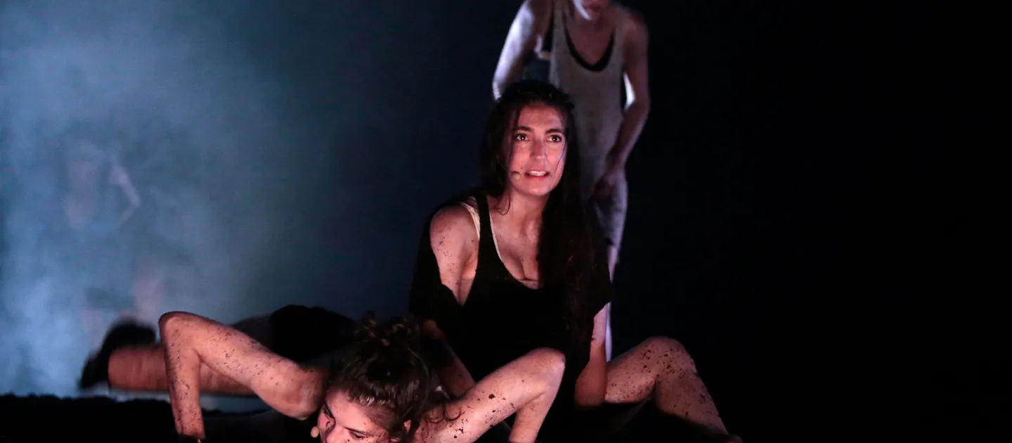 Szene aus "The Wasp Factory", 2013 im Festspielhaus, drei kostümierte Darstellerinnen, eine liegt am Boden mit angewinkelten Armen, eine sitzt dahinter, die dritte steht im Hintergrund, dunkler Bühnenhintergrund mit Nebel