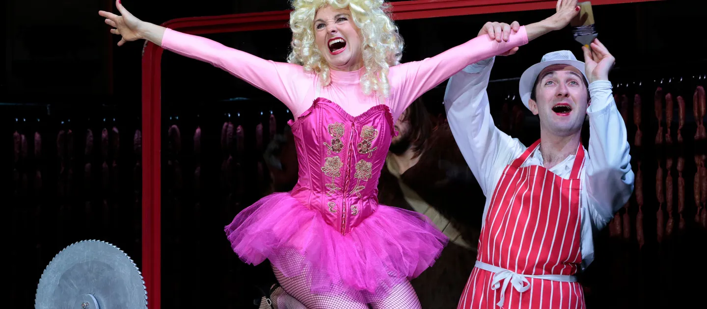 Szene aus "Gloria von Jaxtberg", 2014 im Theater am Kornmarkt, eine Person im pinken Kostüm mit Tutu, blonden Locken und ausgestreckten Armen kniet, rechts hält eine weitere Person einen Arm der knienden Person und trägt einen weißen Hut sowie eine rot-weiß-gestreifte Schürze