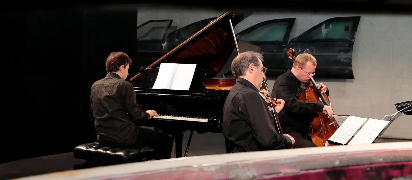 Szene aus "KAZ – Konzerte", 2014 an diversen Spielorten, drei Musiker spielen ihre Instrumente – Klavier, Cello, Violine –, alle in schwarzer Kleidung