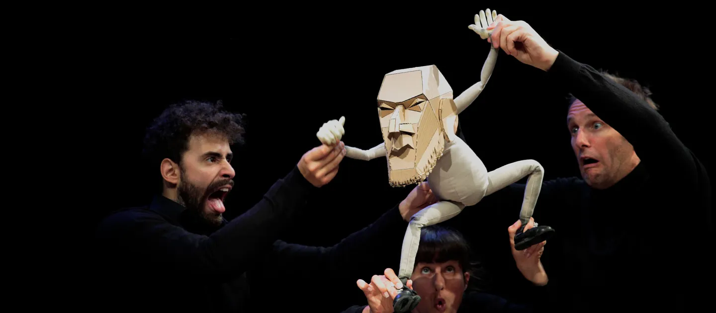Szene aus "The Table", 2014 im Theater Kosmos, drei Personen in schwarzer Kleidung halten eine weiße Puppe in die Luft