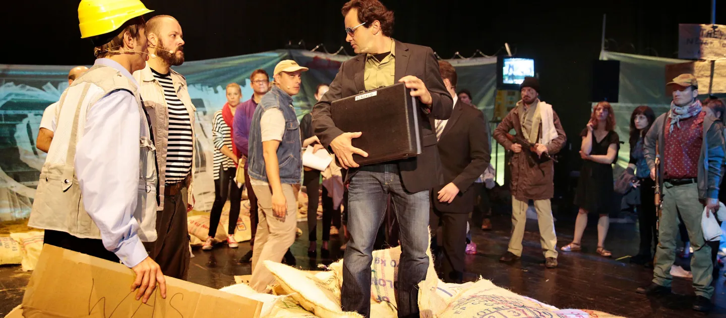 Szene aus "Trans-Maghreb", 2014 im Festspielhaus, drei Personen im Vordergrund – links eine Person mit gelbem Helm und einem Pappkarton in der Hand, mittig ein Mann mit Koffer –, im Hintergrund mehrere Personen
