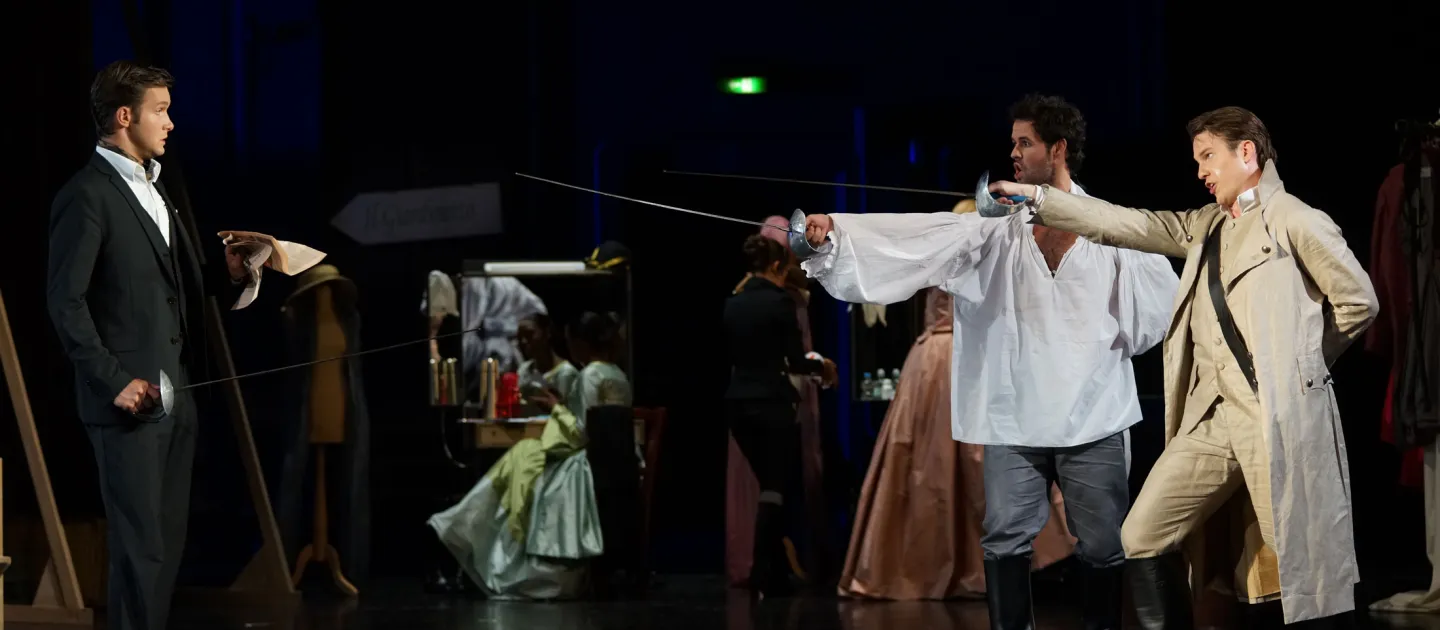 Szene aus "Così fan tutte", 2015 im Theater am Kornmarkt, drei Darsteller im Vordergrund, zwei mit je einem Degen, einer in weißem Hemd und schwarzen Stiefeln, der andere in hellem Mantel, der dritte steht links im schwarzen Anzug, im Hintergrund weitere kostümierte Darstellende