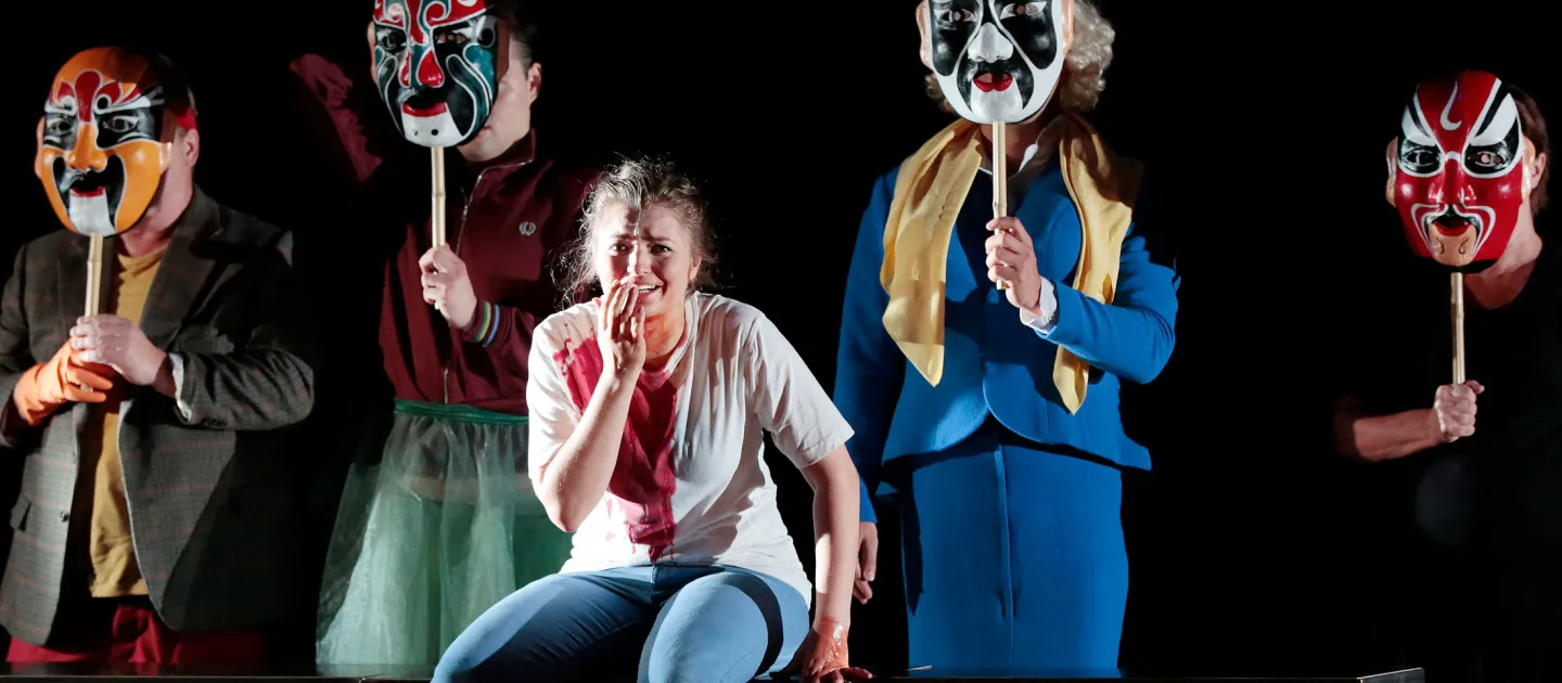 Szene aus "Der goldene Drache", 2015 im Festspielhaus, vier kostümierte Personen halten je eine bunte Maske vor das Gesicht, zentral hockt eine Person und hält ihre linke Hand vor den Mund 